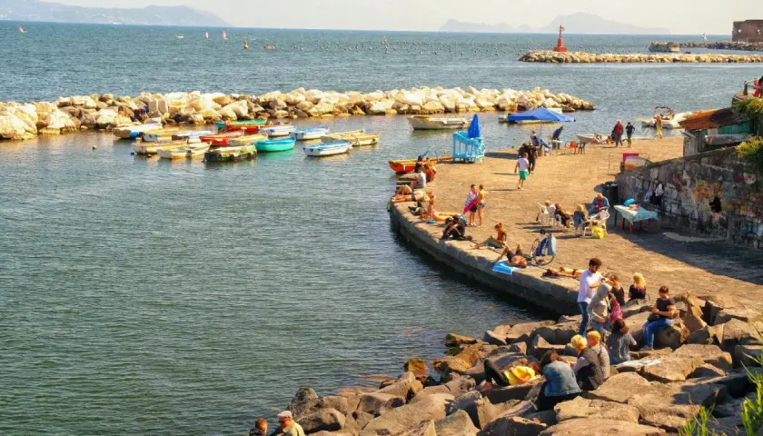 Słoneczna neapol plaża z łodziami i ludźmi odpoczywającymi na kamieniach i molo.
