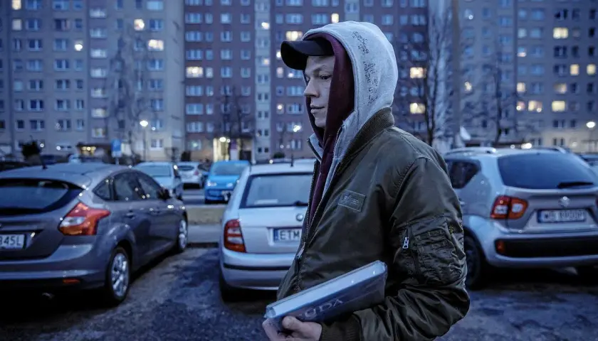 Młody mężczyzna w bluzie z kapturem i kurtce bomberce trzyma w ręku płytę. W tle widać blokowisko i zaparkowane samochody, jakby scena z filmu o innych ludziach.