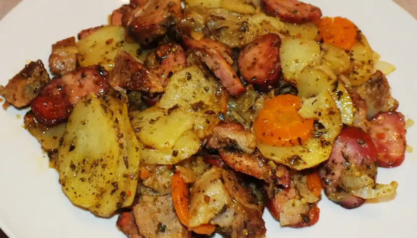 Pyszne dania z ogniska: ziemniaki, kiełbasa i marchewka, doprawione ziołami, podane na białym talerzu.