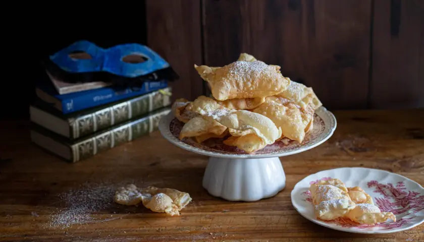 Chiacchiere friabili e bollose, cosparse di zucchero a velo, su un piatto con una maschera veneziana e libri antichi sullo sfondo.