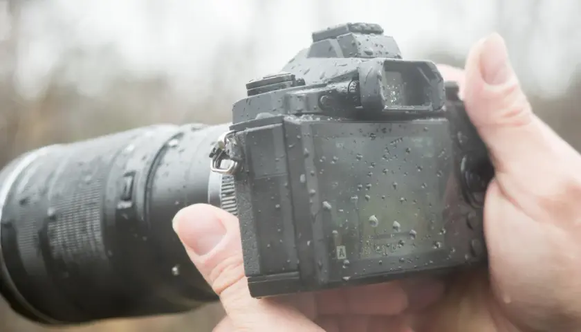 Dłoń trzyma aparat z obiektywem, pokryty kroplami deszczu. To była trudna, ale udana sesja w deszczu.