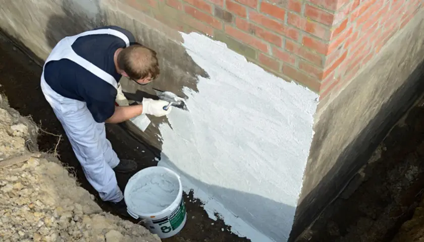 Mann dichtet mit Spachtel und Mörtel die Gartenmauer ab. Ein Eimer mit Abdichtungsmaterial steht bereit.