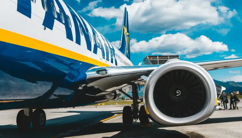 Fragment samolotu Ryanair z widocznym silnikiem i podwoziem na tle błękitnego nieba z chmurami.