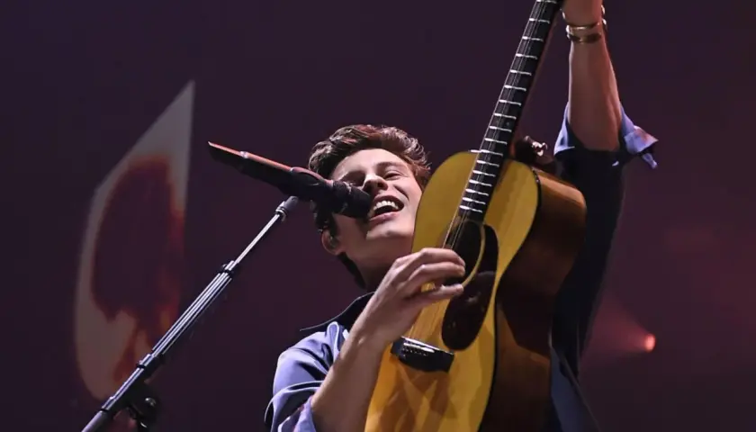 Shawn Mendes na koncercie, śpiewa i gra na gitarze, wznosząc ją w górę.