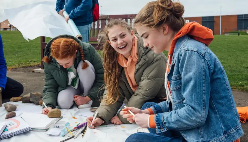 Drei Mädchen sitzen im Freien und malen auf einer großen Papierrolle. Sie lachen und arbeiten zusammen, als ob es eine kleine **Abschlussfeier** wäre.