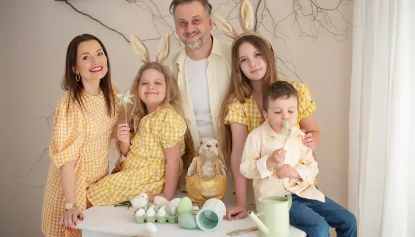 Rodzinna sesja zdjęciowa z zajączkami na głowach. Uśmiechnięci rodzice i dzieci w żółtych strojach pozują na tle wiosennych dekoracji.