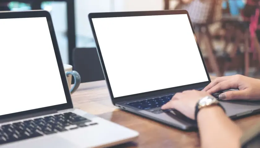 Zwei Laptops auf einem Holztisch. Eine Person nutzt einen Laptop als Monitor, während die Hände auf der Tastatur liegen.