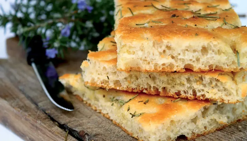 Gestapelte Focaccia-Stücke mit Rosmarin, die an das Backen von Pizza erinnern. Ein Messer liegt daneben auf einem Holzbrett.