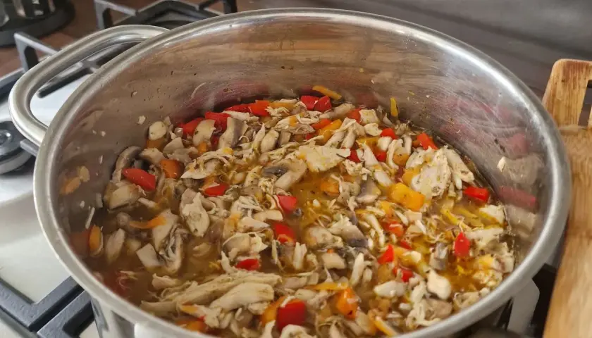 Gęsta zupa z kawałkami mięsa z rosołu, pieczarkami i kolorową papryką w metalowym garnku na kuchence.