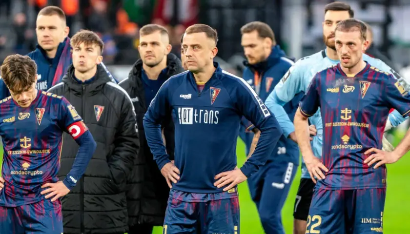 Piłkarze Pogoni Szczecin w ciemnych strojach, z widocznymi herbami klubu. W tle inni zawodnicy i sztab. Wszyscy wyglądają na zamyślonych, być może po trudnym meczu lub w oczekiwaniu na ważne pogoń szczecin - transfery.
