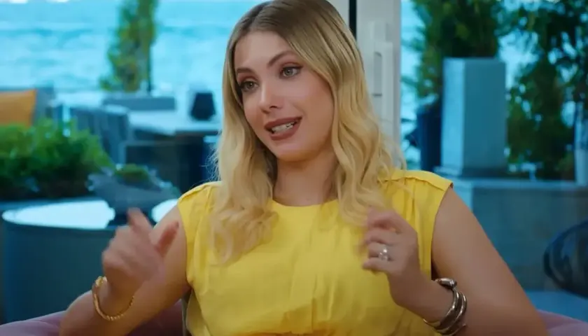 Kobieta w żółtej sukience z wycięciem, gestykulująca rękami, z blond włosami.