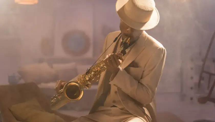 Mężczyzna w eleganckim, beżowym garniturze i kapeluszu gra na saksofonie, emanując prawdziwym jazzowy styl.