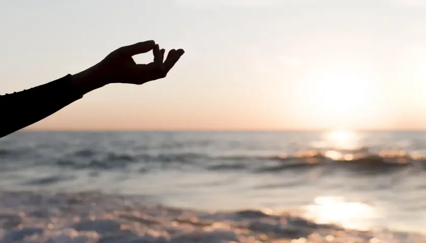 Dłoń w geście mudry na tle zachodzącego słońca nad morzem. Chwila głębokiej mindfulness i spokoju.