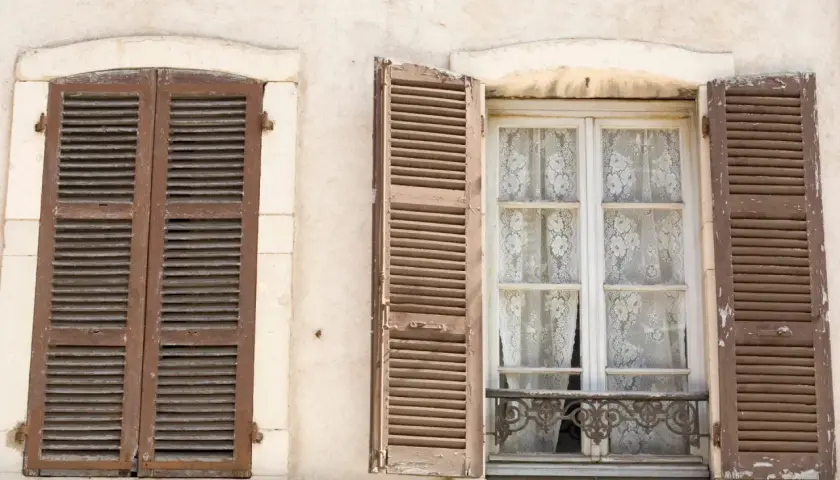 Due finestre d'epoca con persiane in legno marrone. Una è chiusa, l'altra è aperta rivelando tende di pizzo bianco.