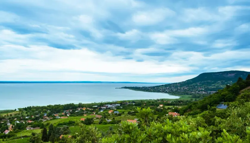 Widok na jezioro Balaton z zielonymi wzgórzami i domkami. Chmury na niebie tworzą dramatyczny krajobraz.