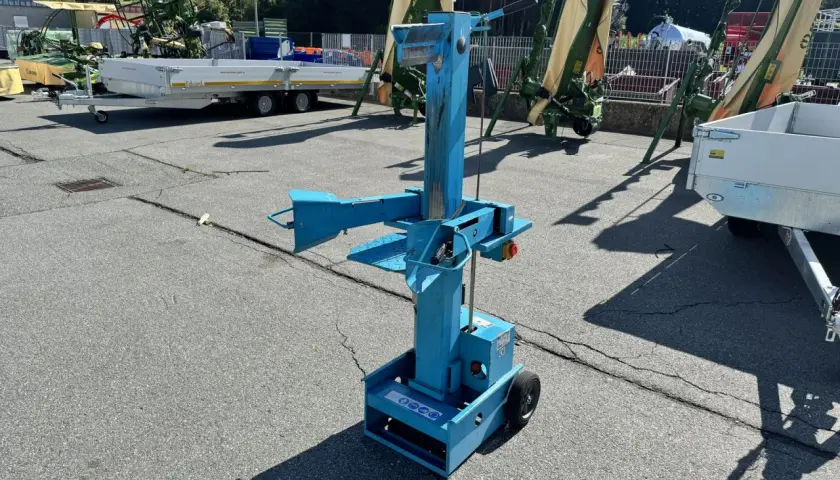 Blauer, gebrauchter Holzspalter mit Rädern steht auf einem Parkplatz. Im Hintergrund landwirtschaftliche Maschinen und Anhänger.