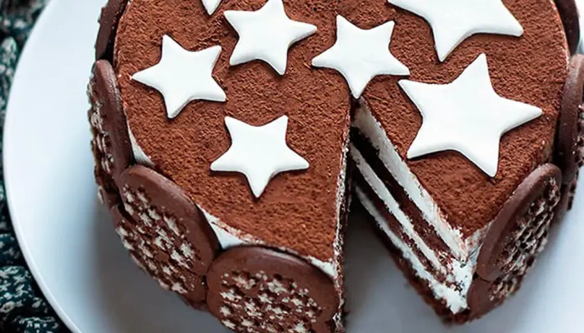 Torta Pan di Stelle ricoperta di cacao, decorata con stelle bianche e biscotti. Una fetta mostra gli strati di crema.