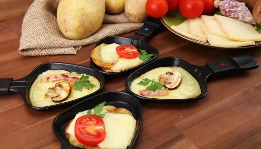 Petites poêles de raclette prêtes à être dégustées, avec pommes de terre, fromage fondu, tomates et champignons. Idéal pour savoir comment couper la raclette.