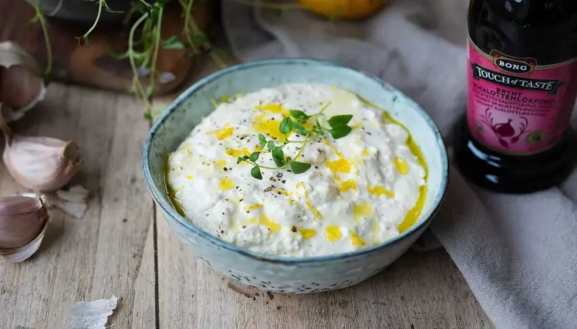 En skål med krämig, vispad fetaost toppad med olivolja och örter. Bredvid syns vitlök, citron och en flaska fond.
