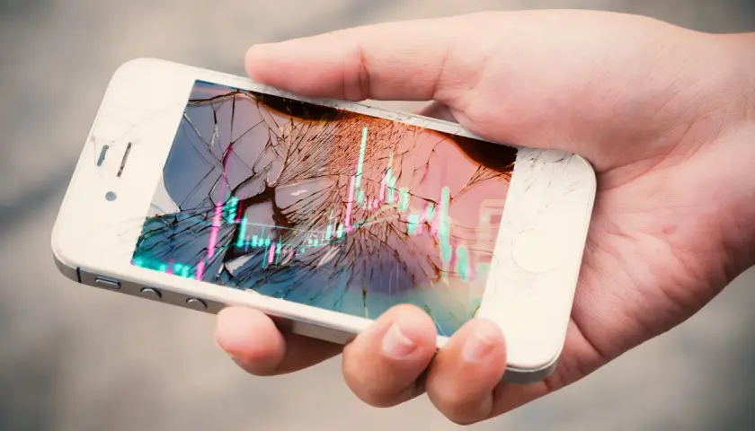 Dłoń trzyma pęknięty smartfon z wykresem giełdowym. Ubezpieczenie ekranu to spokój ducha.