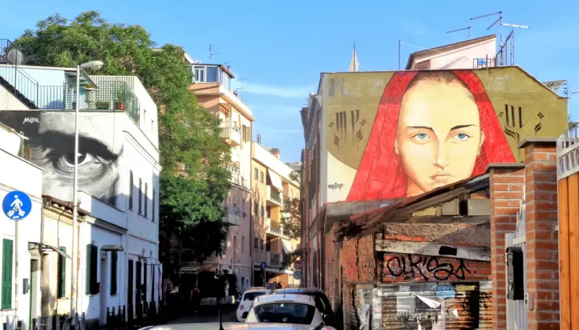 Un grande volto femminile con un velo rosso domina un edificio nel quartiere Pigneto. Accanto, un occhio gigante osserva la strada.