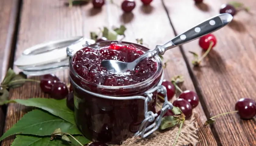 Słoik pełen domowego musu wiśniowego, z łyżeczką gotową do degustacji. Wokół świeże wiśnie i liście.