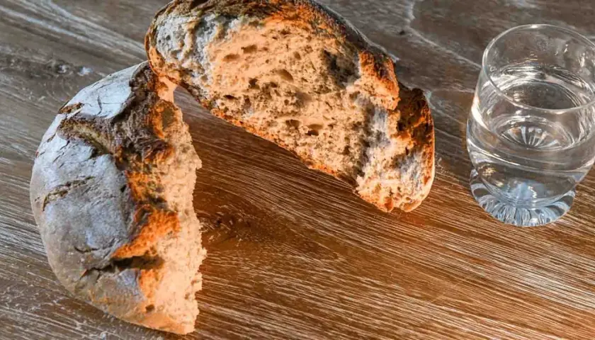 Ein aufgeschnittenes Brot, ein starkes Symbol für Nahrung und Leben, liegt neben einem Glas Wasser auf einem Holztisch.