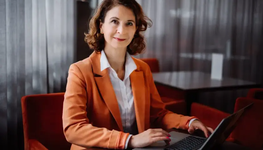 Kobieta w pomarańczowej marynarce pracuje na laptopie w eleganckim wnętrzu.