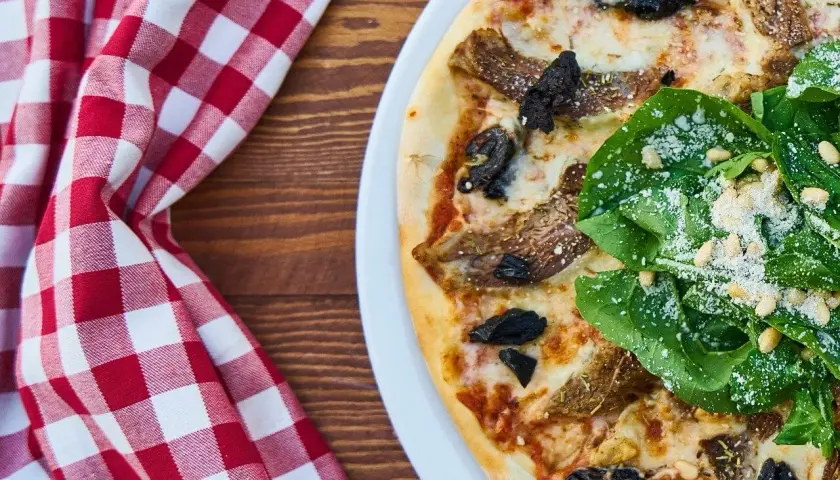 Pizza z rukolą, orzechami pinii i grzybami. Widać kolejność składników na pizzy, od sosu po świeże dodatki.
