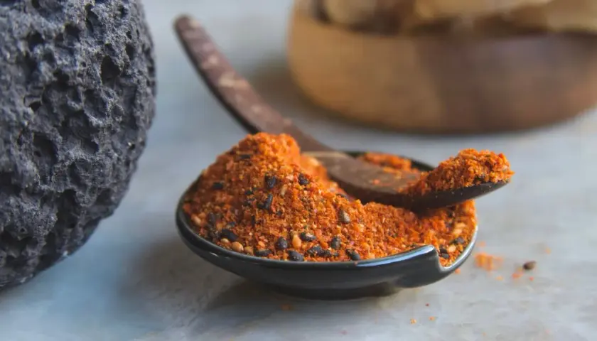 A dark, textured rock sits beside a small bowl filled with a vibrant 7 spice recipe blend. A wooden spoon rests in the bowl, ready to scoop the aromatic mixture.