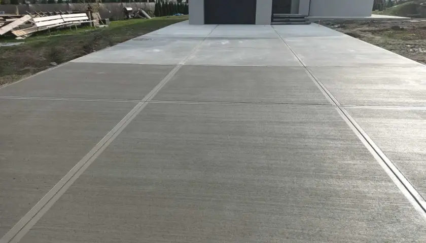 Nowy podjazd z beton stemplowany, z wyraźnymi fugami, prowadzi do nowoczesnego domu z szarą bramą garażową.