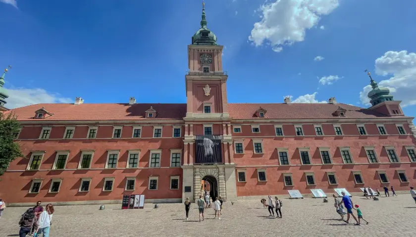 Zamek Królewski w Warszawie, z czerwoną fasadą i wieżą zegarową, na tle błękitnego nieba z chmurami. Plac przed zamkiem tętni życiem.