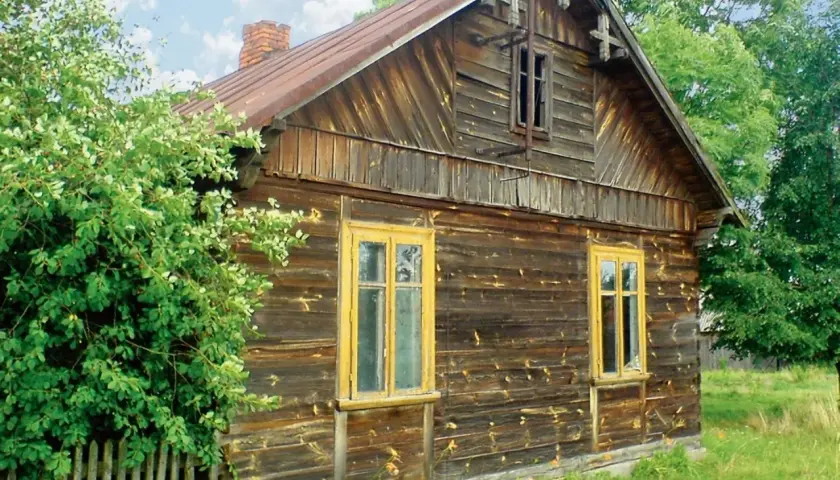 Stary, drewniany dom z żółtymi oknami, otoczony zielenią. Wnętrze tego domu kryje zapewne wiele historii.