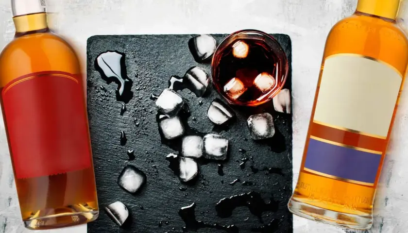 Dwie butelki, jedna z czerwoną etykietą, druga z beżową i fioletową, stoją obok szklanki z lodem i ciemnym płynem. Koniak czy whisky?