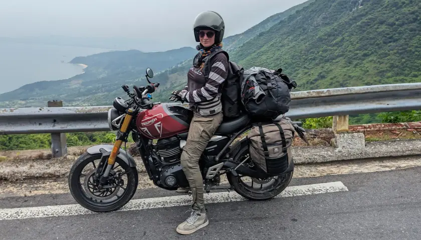 Motociclista con su equipada Triumph Tiger 400, lista para la aventura en una carretera con vistas al mar y montañas.