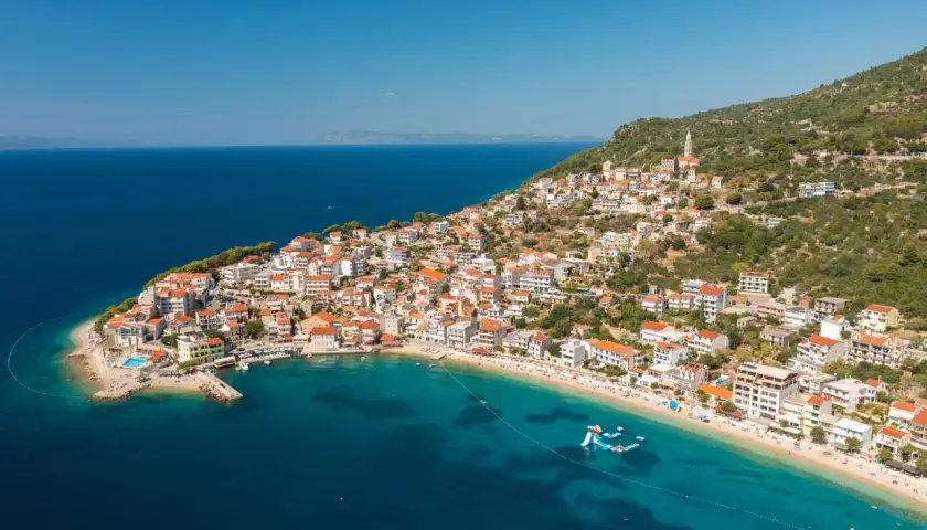 A picturesque village clings to a hillside on the Balkan coast, overlooking a turquoise bay with a sandy beach and inflatable water slides.