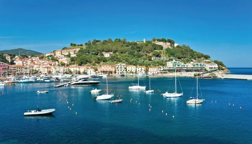 Vue panoramique de Porto Azzurro, avec ses maisons colorées, sa colline verdoyante et son port animé rempli de voiliers et de bateaux.