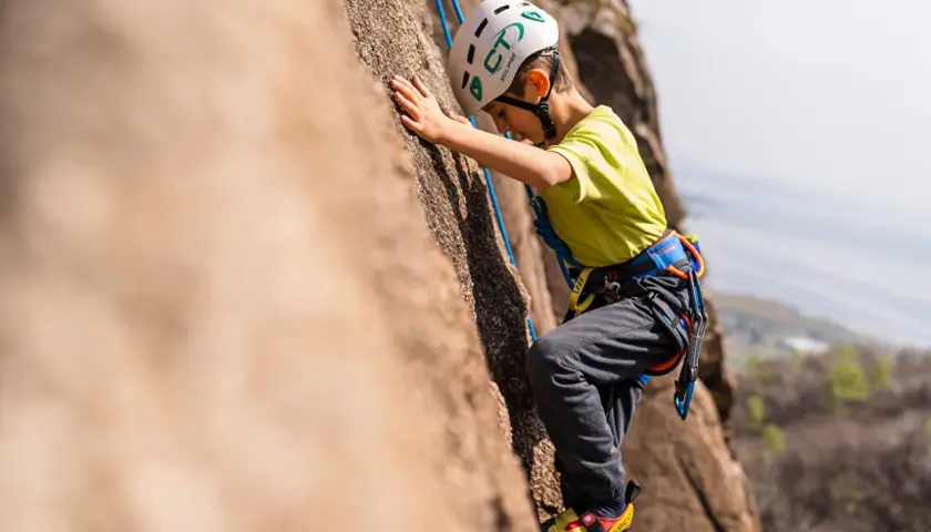 Bambino con casco e imbracatura si arrampica su una parete rocciosa. Un'avventura di arrampicata per bambini, anche se ne avesse 3 anni!