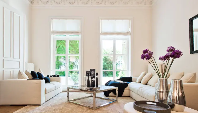Helles Wohnzimmer im Hamptons-Stil mit beigen Sofas, einem Chrom-Couchtisch und großen Fenstern, die ins Grüne blicken.