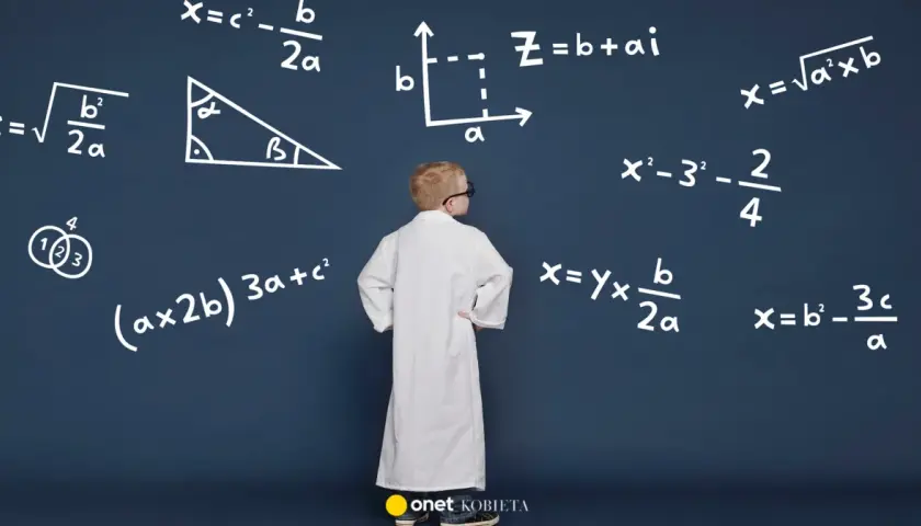 Chłopiec w białym fartuchu stoi przed tablicą z wzorami, w tym wzorem na przyspieszenie.