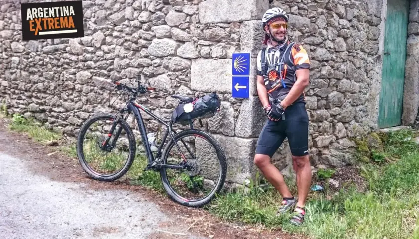 Ciclista sonriente junto a su bici en un camino primitivo, con señal de flecha y concha de peregrino.