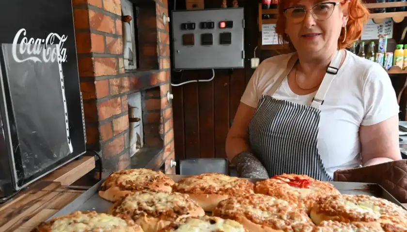 Kobieta z rudymi włosami prezentuje świeżo upieczone pizze w najstarszej pizzerii w Polsce. W tle widać ceglany piec i tablicę z logo Coca-Coli.