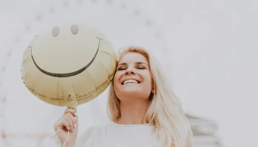 Uśmiechnięta blondynka trzymająca balon w kształcie buźki. Dobre żarty po angielsku poprawiają humor.