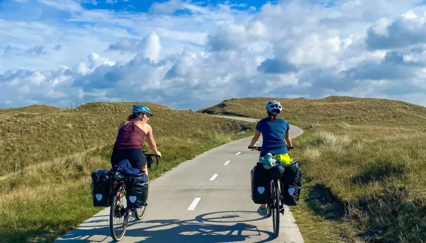 Two cyclists with panniers enjoy a scenic path through grassy dunes, perfect for Netherlands bike trips.