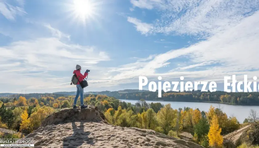 Jesień nad jeziorem. Kobieta z aparatem podziwia krajobraz, szukając ełk atrakcji dla dorosłych. Słońce, chmury i kolorowe drzewa tworzą malowniczy widok.