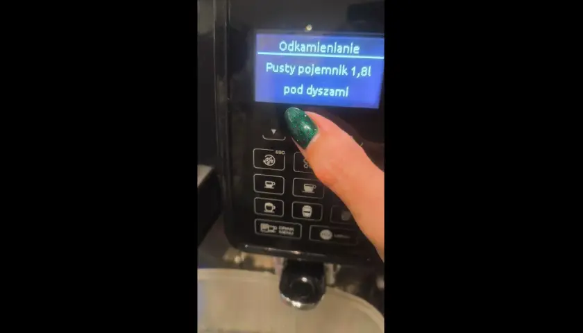 Palec z zielonym lakierem naciska przycisk na ekspresie DeLonghi. Na ekranie wyświetla się komunikat "Odkamienianie. Pusty pojemnik 1,8l pod dyszami".