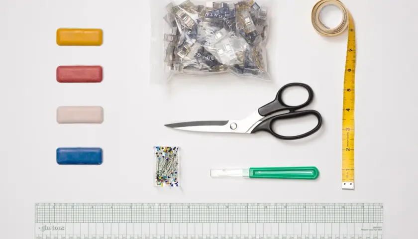 A collection of essential sewing tools, including colorful clips, pins, scissors, a measuring tape, a seam ripper, and a ruler, are laid out on a white surface.