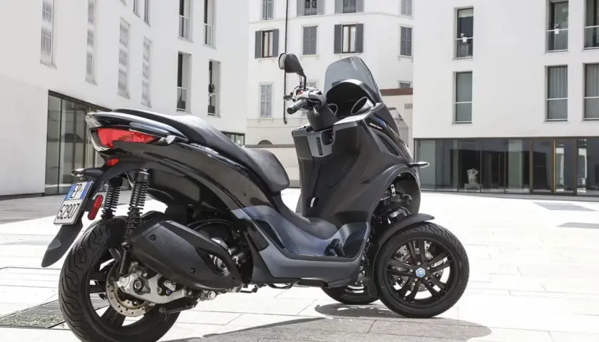 Un Piaggio MP3 300 negro aparcado en una plaza urbana. Su diseño de tres ruedas ofrece estabilidad y un consumo eficiente.