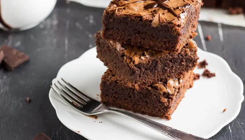 Pyszne ciasto dla odchudzających się: trzy czekoladowe brownie ułożone na talerzyku, obok widelec i kawałki gorzkiej czekolady.