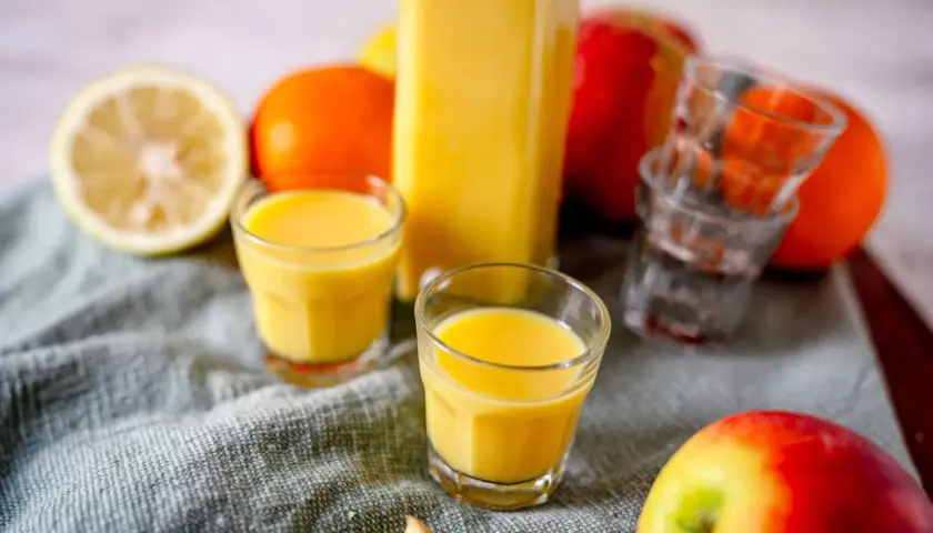 Frische Zutaten für Ingwer Shots selber machen: Zitrone, Orangen, Äpfel und ein Glas voller goldgelbem Shot.